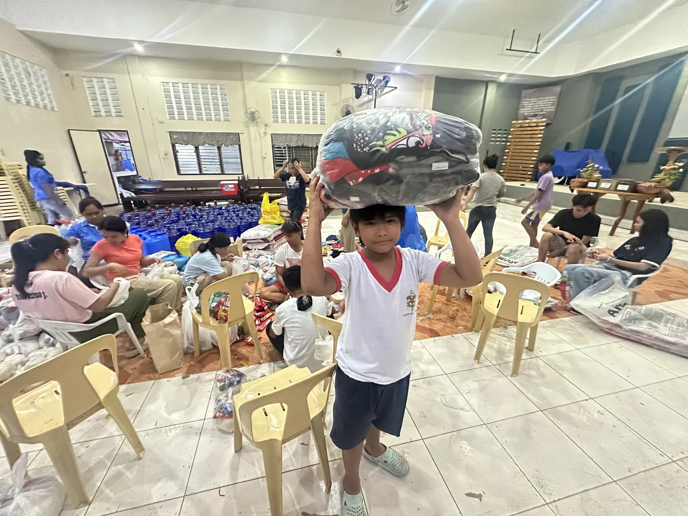 A child carrying a packed bag among adults preparing supplies